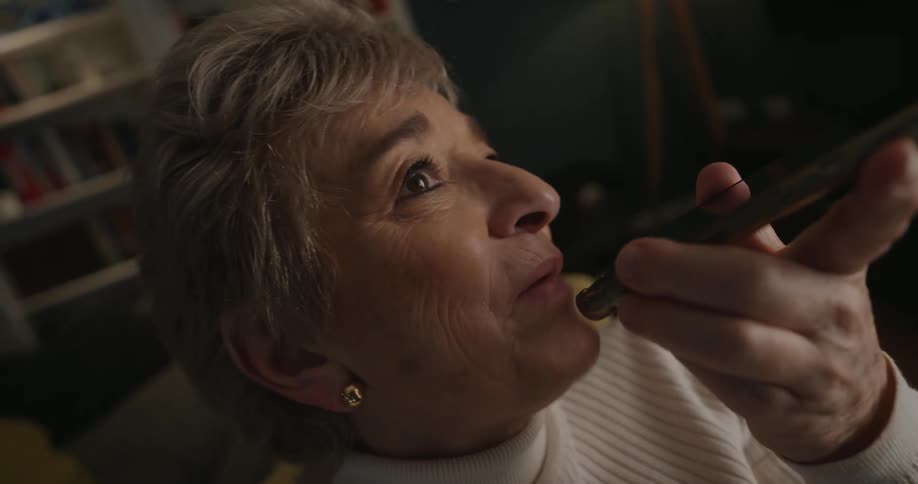 A very closeup shot focuses on the profile of an elderly woman who is smiling and speaking into her smartphone which she holds near her mouth while speaking