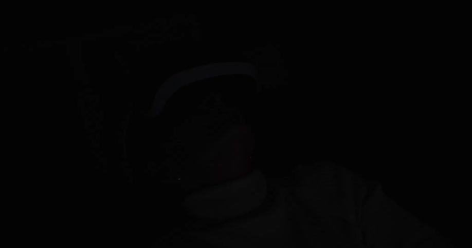 A lowangle shot shows a woman wearing a white turtleneck sweater and a virtual reality vr headset she is sitting in a room that appears to be a living area with a bookshelf and a lamp visible in the background mostly in shadow except when the headset
