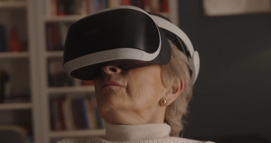An elderly woman wears a virtual reality headset and a white turtleneck sweater looking forward with a serious expression against an indoor background of a blurred bookshelf