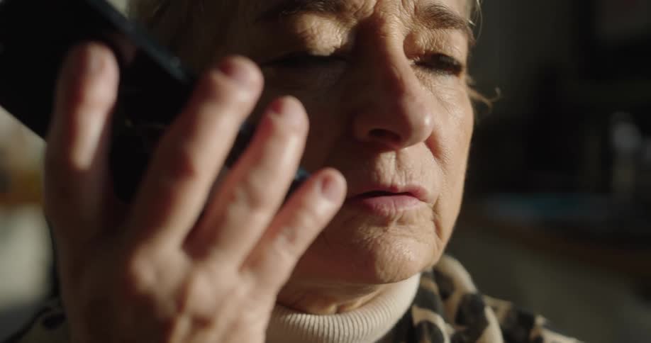 The video shows a tight closeup of an elderly womans face as she holds a dark smartphone up to her ear and speakssunlight illuminates the side of her face highlighting her worried or concerned expression as she listens and responds to the call