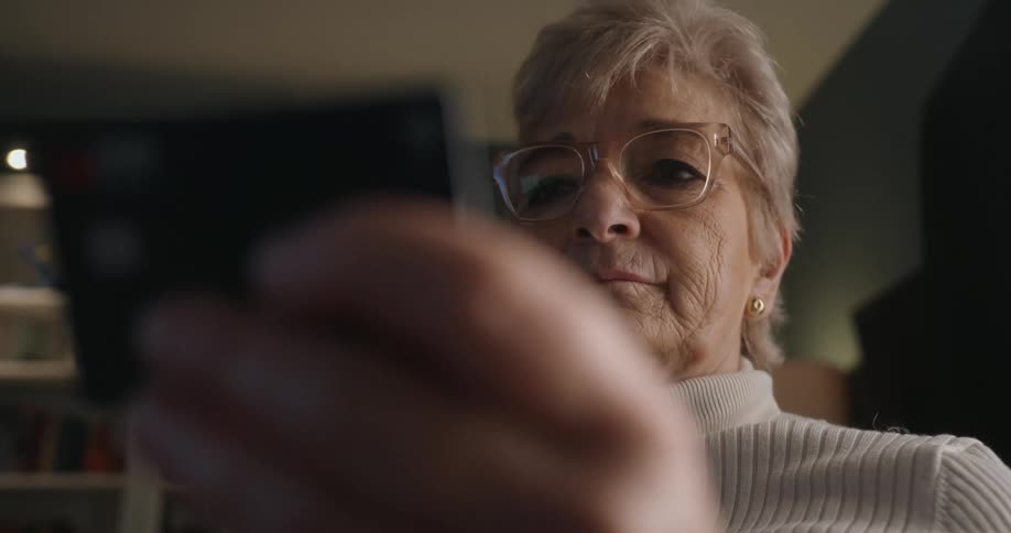 The video clip is a closeup of an elderly woman wearing glasses and a lightcolored turtleneck sweater she is looking intently at something she is holding in the foreground which is mostly out of focus possibly a card or small device