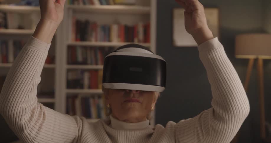 An elderly woman raises her arms and hands in the air over her head as she wear a virtual reality set in a living room