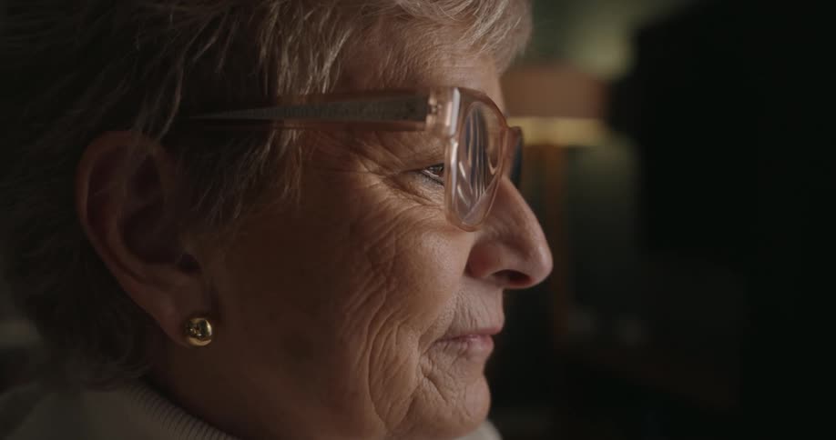 The profile of an elderly woman wearing glasses looking intently toward the right with a neutral focused expression while sitting in a dimly lit room with a bookcase and lamp visible in the background