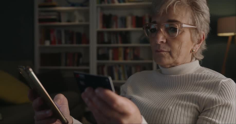 An elderly woman wearing a white turtleneck sweater and large eyeglasses shows her intently looking back and forth between a smartphone she holds in one hand and a dark credit card in the other with a book shelf behind her in an indoor setting