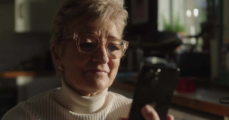 An elderly woman wearing largeframed glasses and a white turtleneck sweater holds a dark smartphone in her hands and smile while sitting in an indoor naturally lit environment