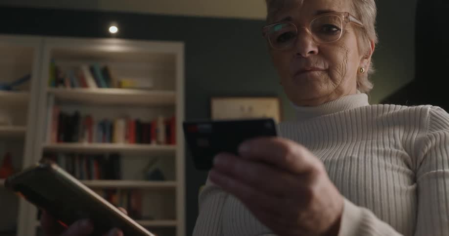 An elderly woman wearing glasses and a white turtleneck sweater is shown holding a smartphone in one hand and a black credit card in the other concentrating on performing a transaction with a brightly lit bookshelf visible in the dark background