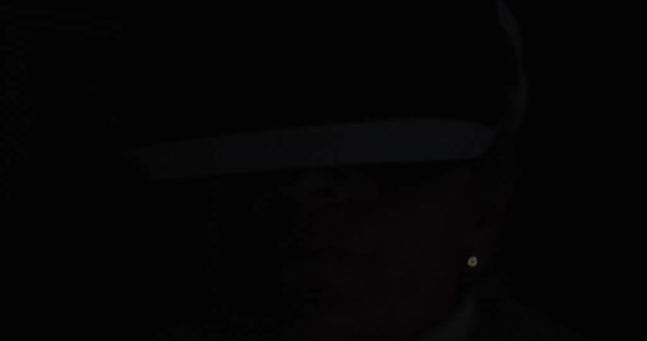 This is a closeup video primarily in darkness showing the profile of a persons head wearing a white and black virtual reality vr headsetthe vr headset briefly rotates upward revealing the persons mouth and chin before the scene fades back into