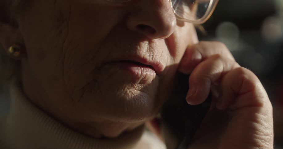 An extreme closeup shot focuses on an elderly woman who is talking on a smartphone with her lips moving as she speaks while a hand holds the phone near her cheek