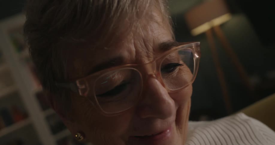 An elderly woman wearing large clearframed glasses is captured in an extremely close lowangle shot laughing set against a blurred lowlit indoor background showing bookshelves and a lamp