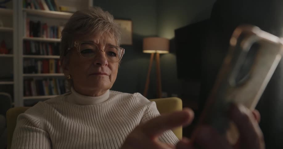 An older woman sits in a cozy living room typing on a smartphone