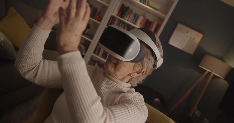 An older woman in a white turtleneck sweater sits in a chair wearing a virtual reality headset and moves her hands in front of her in a dimly lit living room with a bright bookshelf