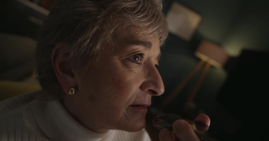 An older woman is sitting in a cozy living room with her smart phone held to her mouth talking into the microphone