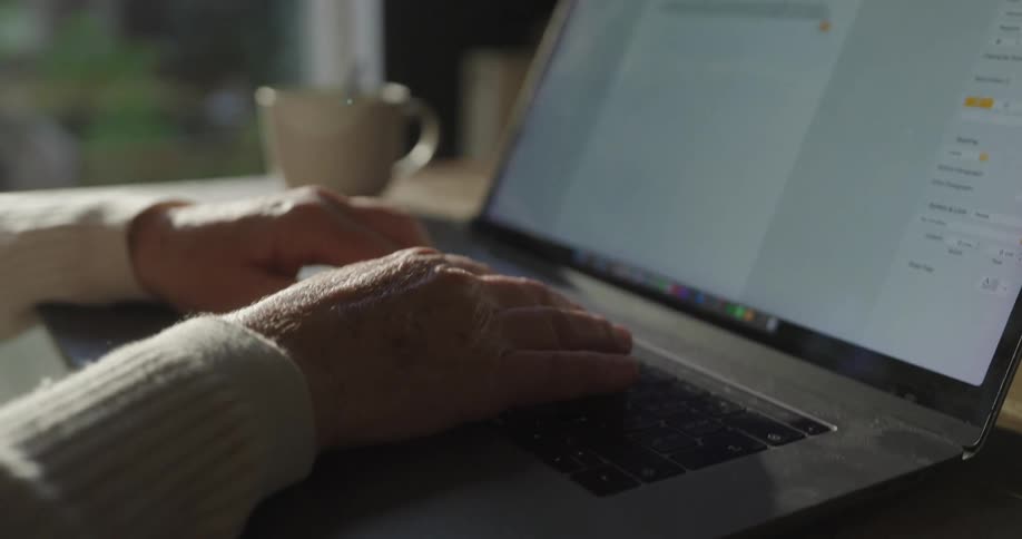 An elderly female sits in a sunlit room typing on a laptop next to her is a mug and spoon