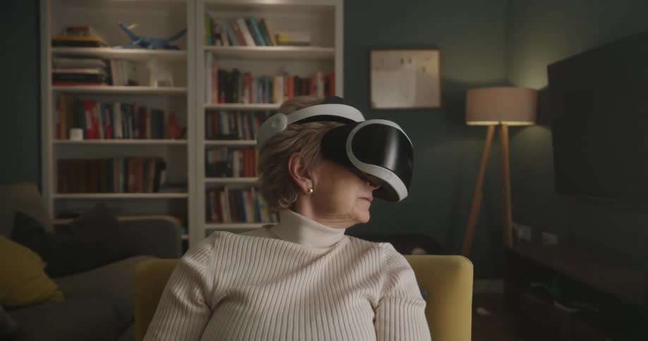 An older woman sits in a chair wearing a virtual reality headset moving her head slowly and engaging with the immersive experience in a brightly lit cozy living room
