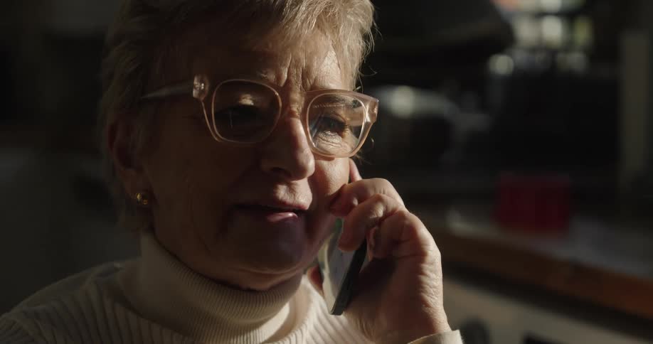 An elderly woman wearing prominent glasses and a white turtleneck sweater shows her speaking into a dark smartphone held to her ear in a dimly lit indoor environment that is partially illuminated by a bright shaft of natural light