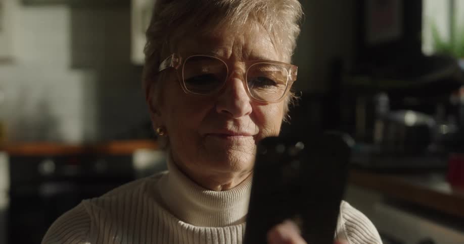 A closeup shot of an elderly woman wearing eyeglasses and a lightcolored turtleneck sweater shows her holding a dark smartphone device and looking at its screen