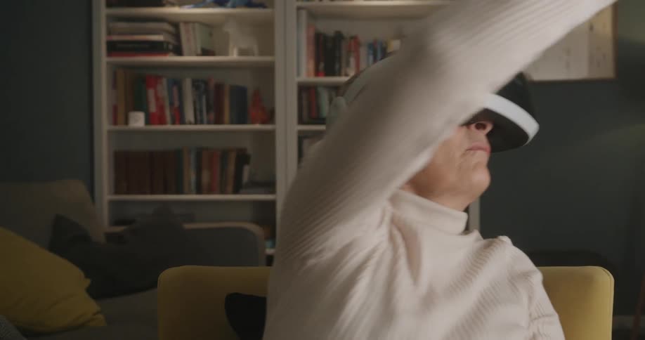 The video features an older woman wearing a white turtleneck sweater and a virtual reality vr headset sitting in a chair in a comfortable living room settingshe is actively moving her arms up down and out to the sides in expressive gestures sugg