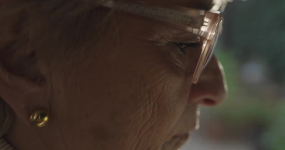 A closeup profile of an elderly woman wearing glasses and a gold earring with a neutral expression looking forward in a dimly lit indoor setting