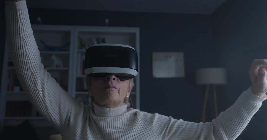An elderly woman wearing a white turtleneck sweater and a virtual reality headset is seen sitting indoors actively moving her arms and hands as she interacts with the unseen virtual environment with a bookshelf visible in the background