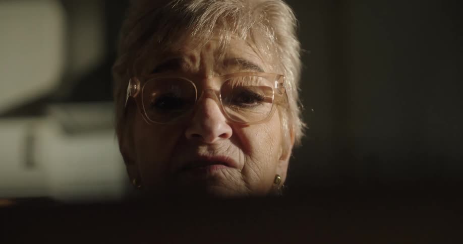 An elderly woman with short white hair and clearframed glasses looks downward with a focused expression in a closeup dimly lit by a source above and behind a dark foreground surface