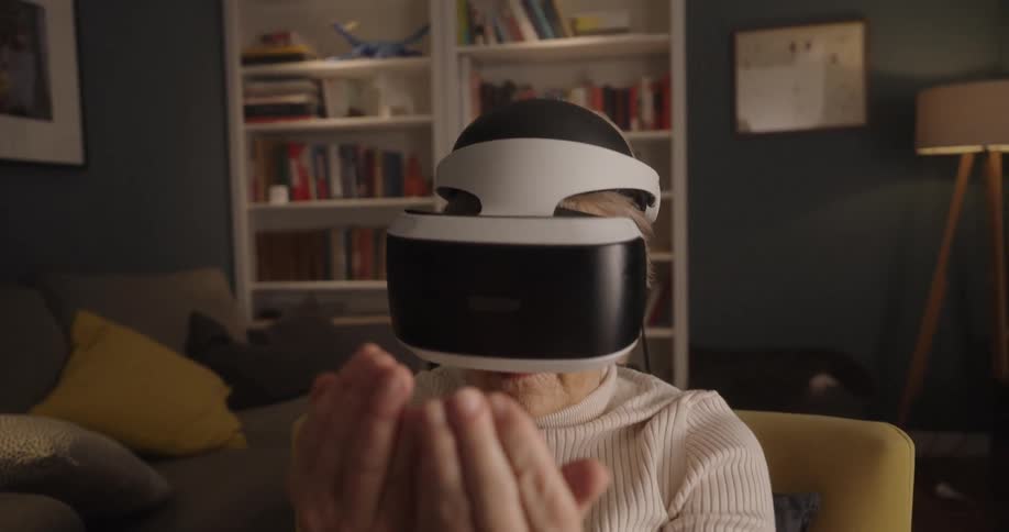 An elderly woman seated in a yellow armchair wears a virtual reality headset and raises and moves her hands in front of her face as if interacting with objects in a digital space in a living room furnished with a large bookshelf