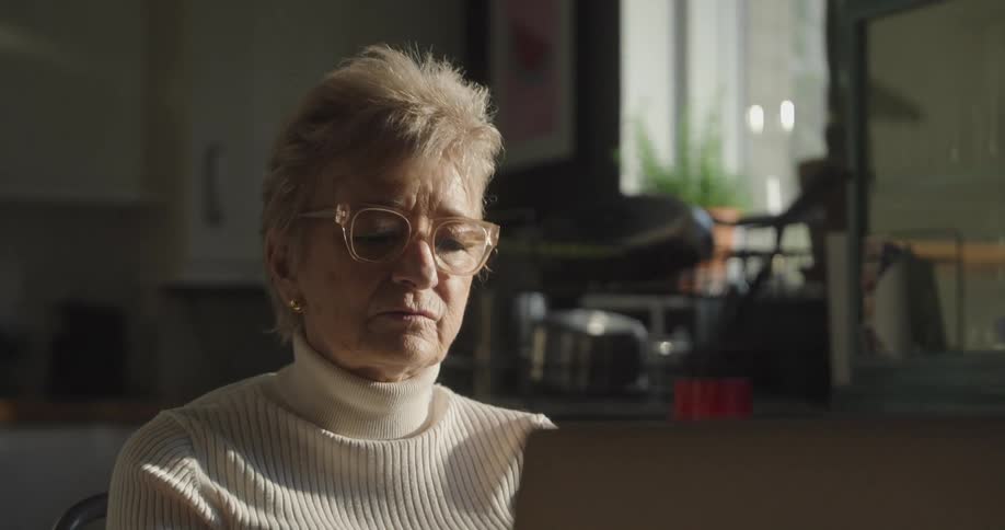 A close view of an older woman in a white turtleneck and glasses talking during a video call on a laptop in a bright kitchen setting