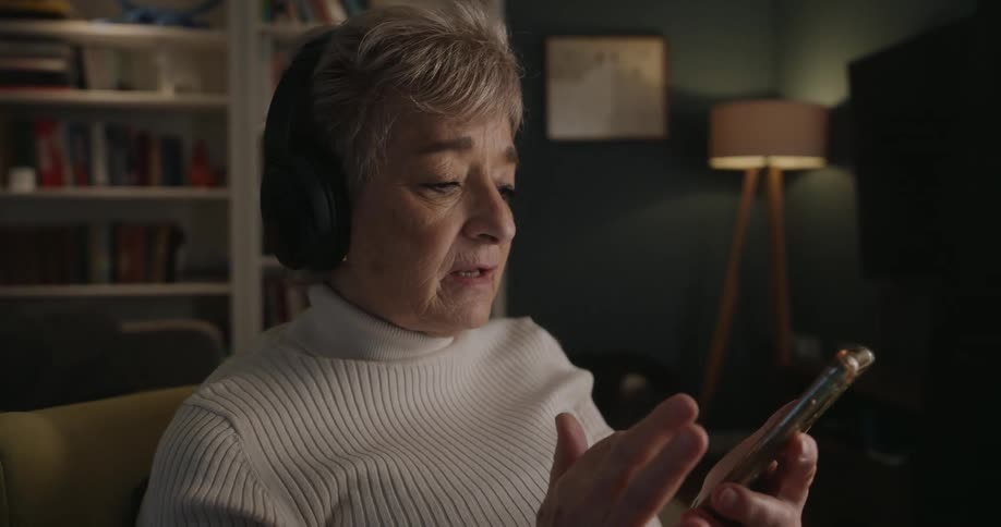 An elderly woman is sitting in a living room with head phones on singing while scrolling on a smartphone