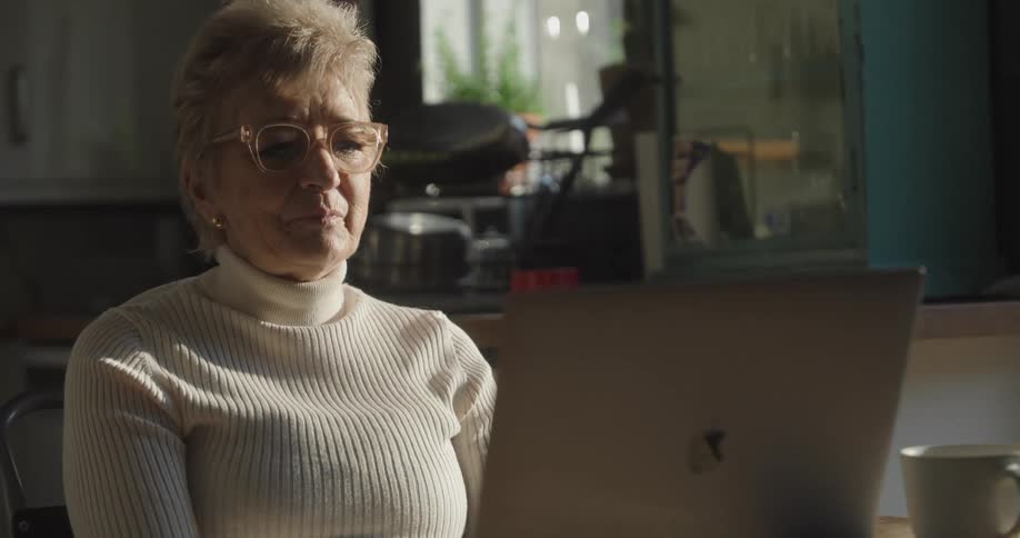 An elderly woman wearing a white turtleneck sweater and large eyeglasses is seated indoors speaking actively into her laptop lluminated by bright natural light coming from the background