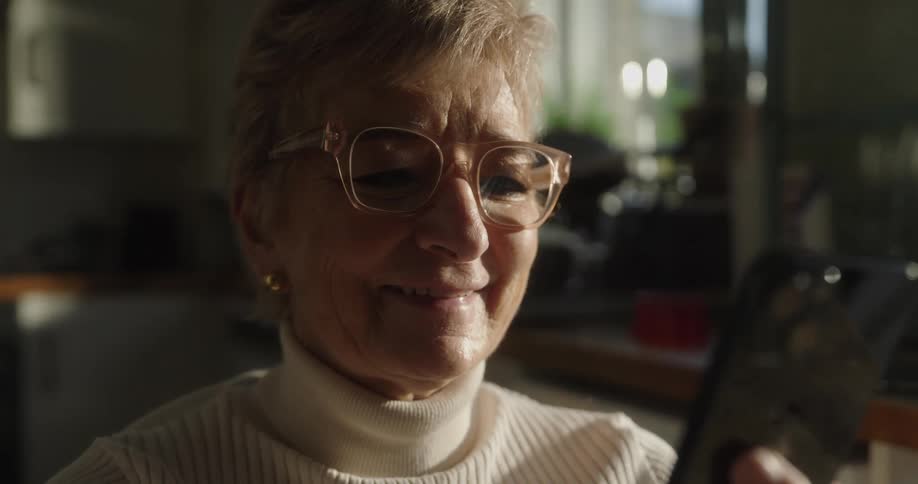 An elderly woman in a white turtleneck and glasses smiles while looking at her smartphone in warm indoor lighting