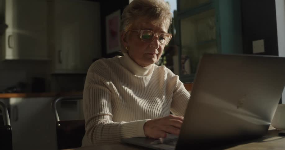 A close view of an older woman in a white turtleneck sweater and glasses sitting at a table and typing on a silver laptop in a sunny kitchen