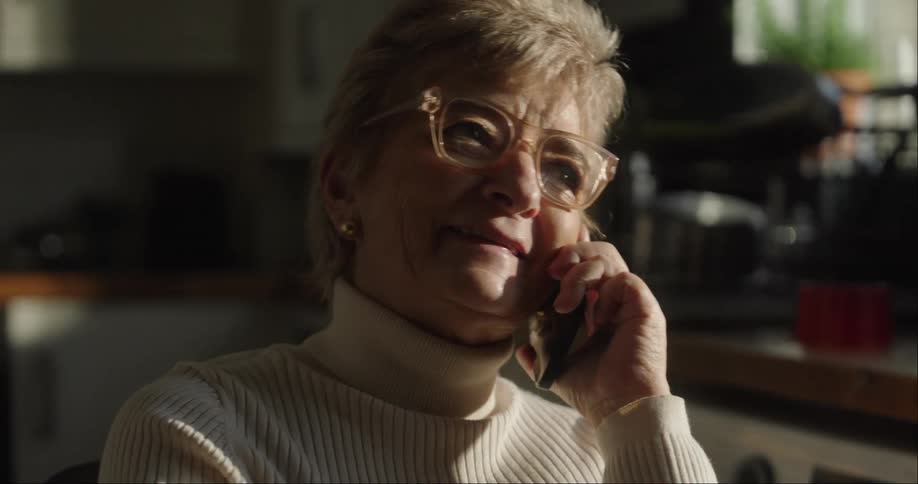 An elderly woman in a white turtleneck and glasses smiles while talking on a cellphone in a dimly lit indoor space with sunlight hitting her face