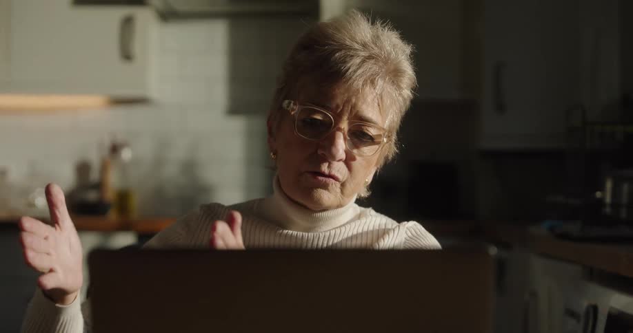 The video features an elderly woman with short blonde hair and glasses wearing a white turtleneck sweater sitting in a dimly lit kitchen and actively video chatting on a laptopshe uses expressive hand gestures raising her hands and arms possibly
