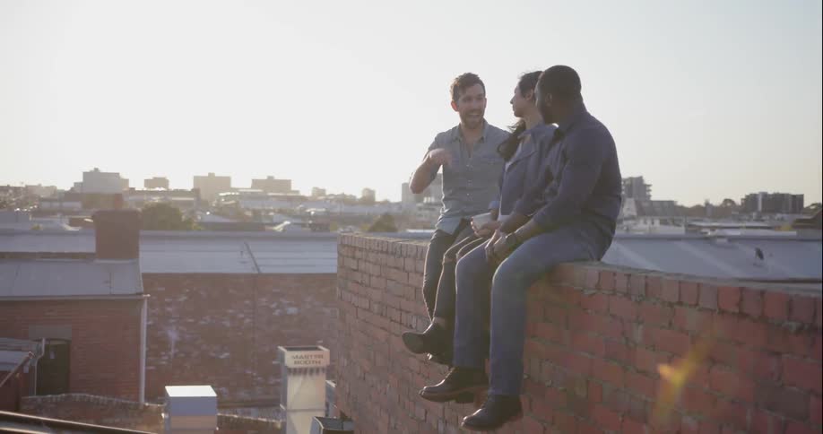 Two men and a woman sit on a brick ledge on a rooftop