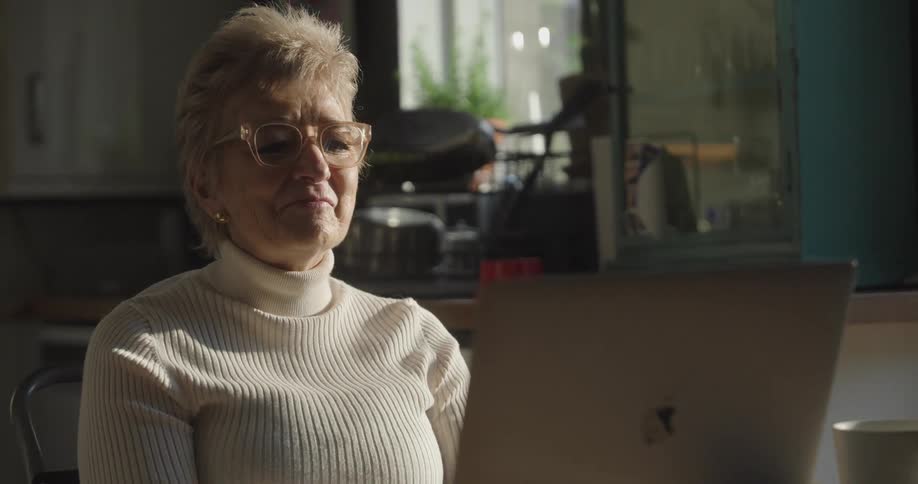 Adult woman wearing glasses talking and laughing in front of a laptop on a kitchen indoors during day time