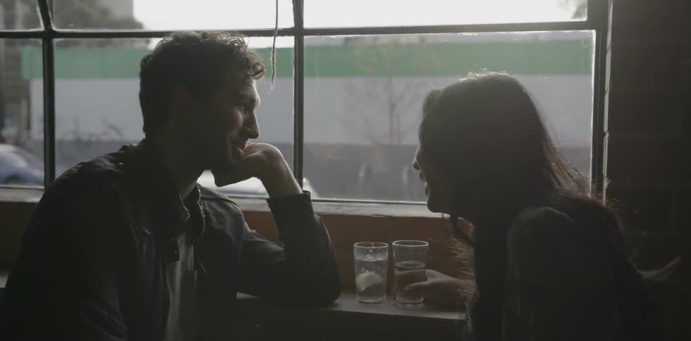A man at a woman sit by a window with glasses of water talking and smiling