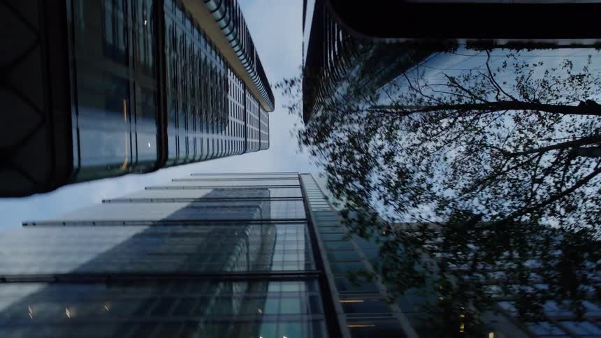 Several towering glass skyscrapers and their reflections making the surrounding architecture feel immense against the backdrop of a partially cloudy sky and surrounding tree branches