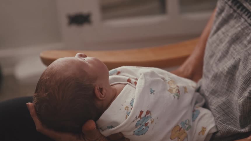 A newborn baby wrapped tightly in a patterned swaddling blanket resting securely in the cradling hands of a caregiver who is partially visible wearing dark pants and a patterned textile while sitting in a wooden rocking chair
