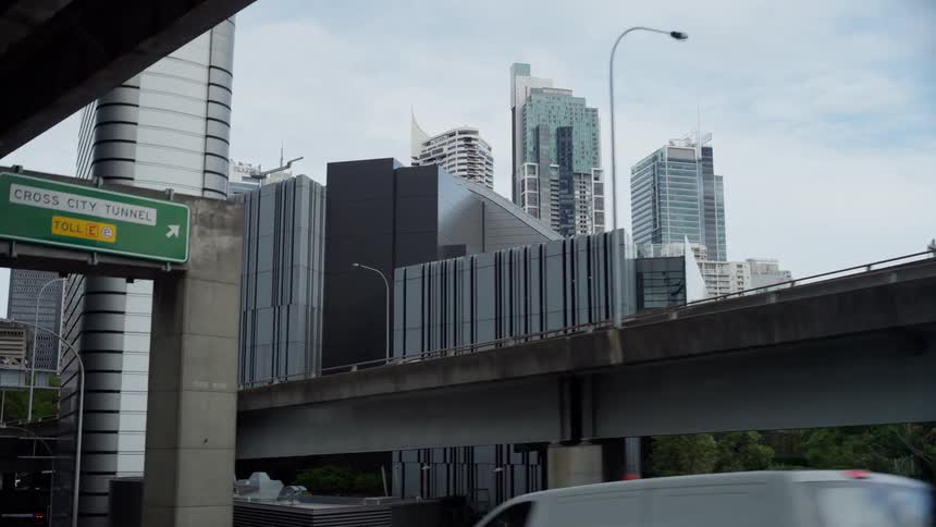 A view from below a highway shows cars driving past a toll sign and several modern skyscrapers and overpasses in a dense urban area