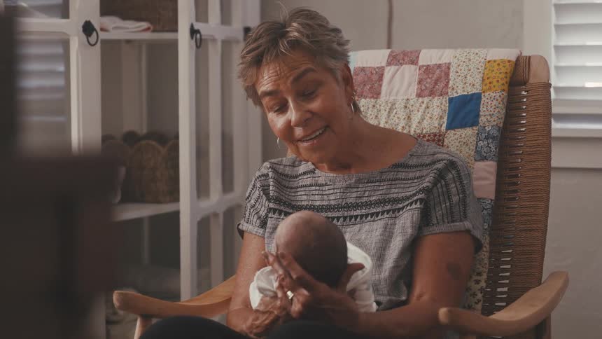 An older woman sits in a rocking chair tenderly cradling a newborn baby in her arms