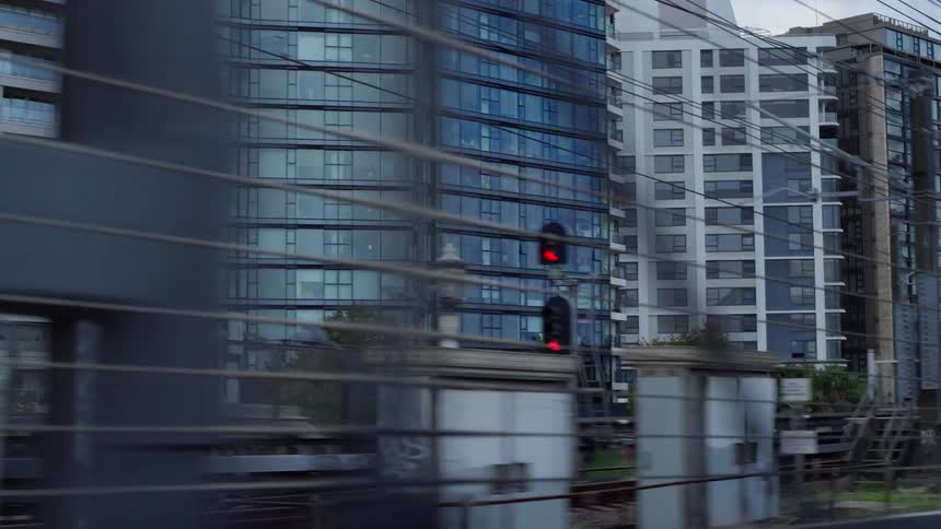 A side view from a moving train shows commercial and residential buildings with reflective glass windows as a yellow train quickly passes on an elevated track