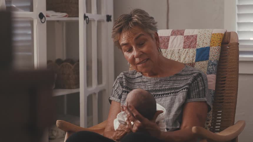 An older woman with short grey hair is sitting in a rocking chair with a colorful patchwork quilt draped over it gently holding and looking down at a swaddled newborn baby the scene is bright and indoors suggesting a cozy domestic setting