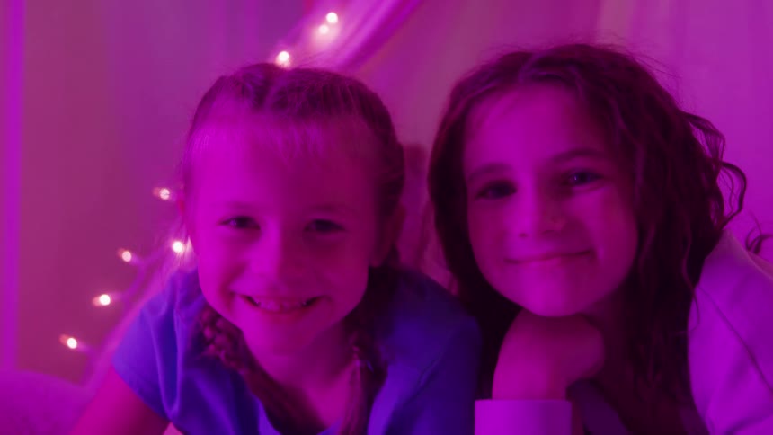 Two smiling kids lie on a bed under purple string lights looking forward happily