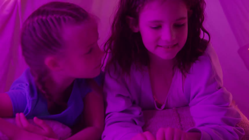 Two young girls illuminated by a vibrant pink light lie close together happily reading a book and occasionally looking up at each other and smiling inside a cozy tentlike fort