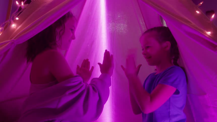 Two young girls are inside a blanket fort or teepee decorated with string lights playing a clapping game and smiling the interior of the fort is brightly lit with a vertical beam of intense pinkpurple light