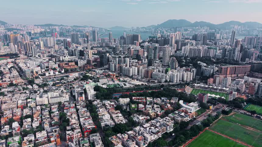 An aerial shot captures an expansive dense city with highrise buildings filling the foreground and midground leading back toward a harbor area and large mountains under a slightly hazy sky