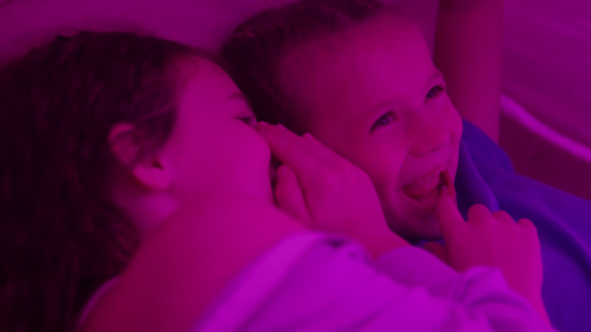 Two girls are sharing a secret while lying down under warm colored light as the girl on the right reacts to the whisper with a wideopen mouth and laughter