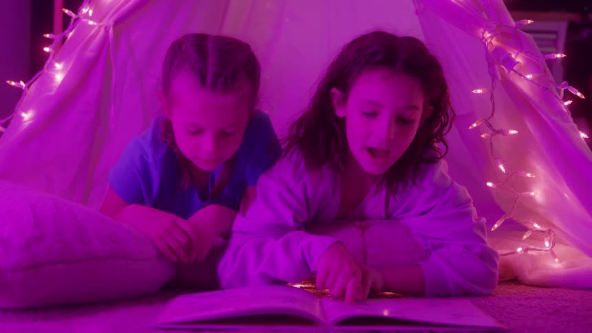 Two young girls are lying on their stomachs inside a teepee tent decorated with string lights reading a book together under pink and purple ambient lighting