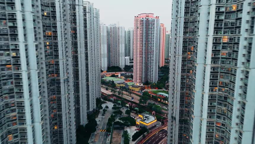 Moving through a canyon of towering residential skyscrapers the view reveals a busy street below where warm window lights begin to flicker on against the cool evening sky