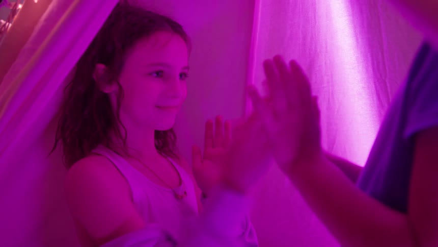 The video captures two children sitting inside a blanket fort or tent illuminated by bright dramatic magenta and purple lightingthe focus is on one smiling girl as she plays a handclapping game with the other child whose back and raised hands are v