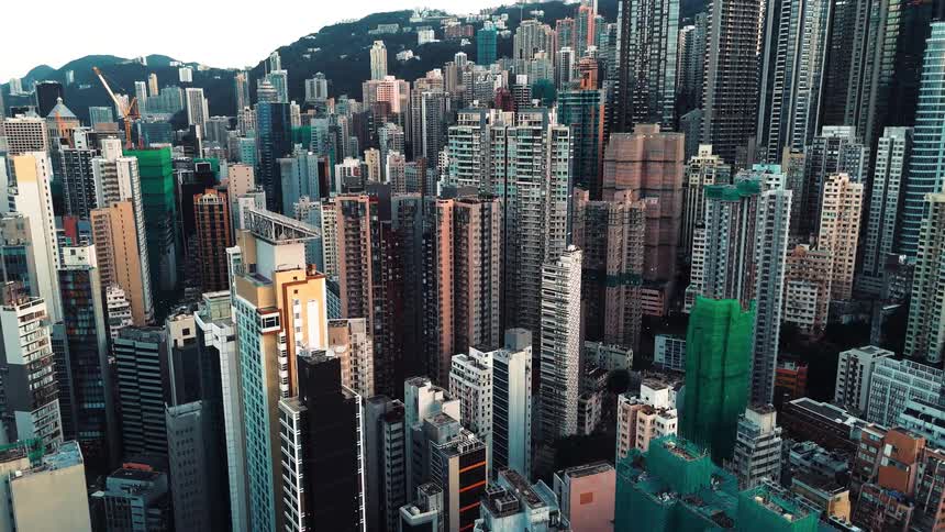 An aerial slow zoom out shot shows a densely packed urban cityscape of numerous tall residential and commercial buildings clustered against a backdrop of green hills